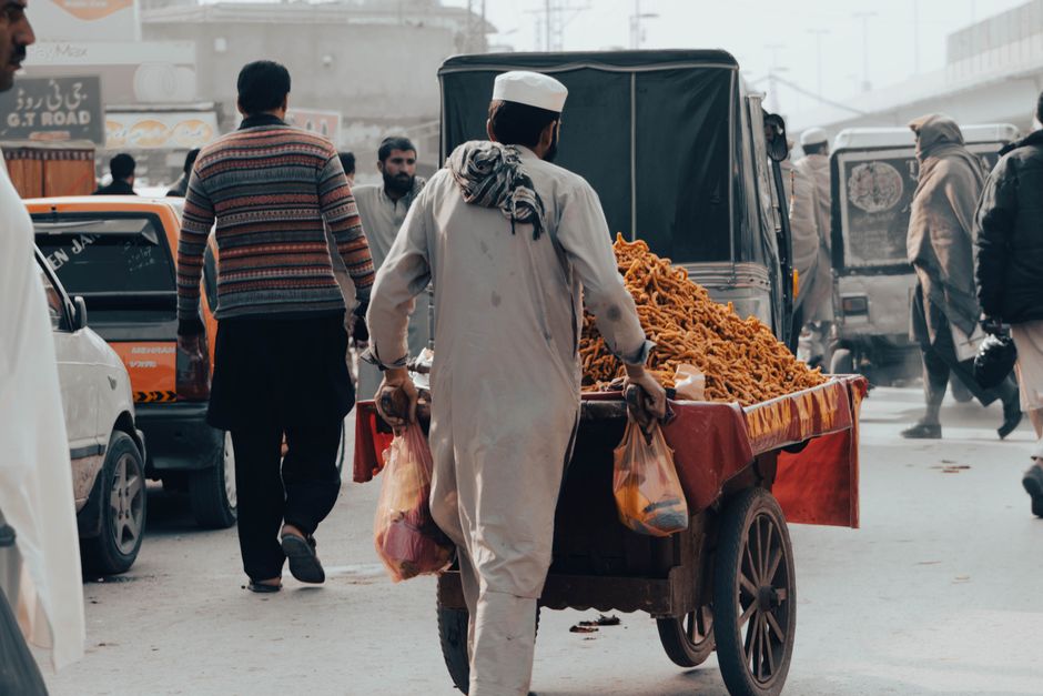 A vendor pushes a street food cart through a bustling urban market street with vehicles and people.