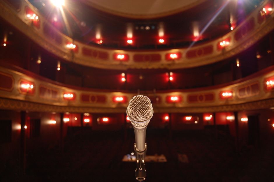 A microphone stands on a theater stage with ornate balconies and warm lighting.