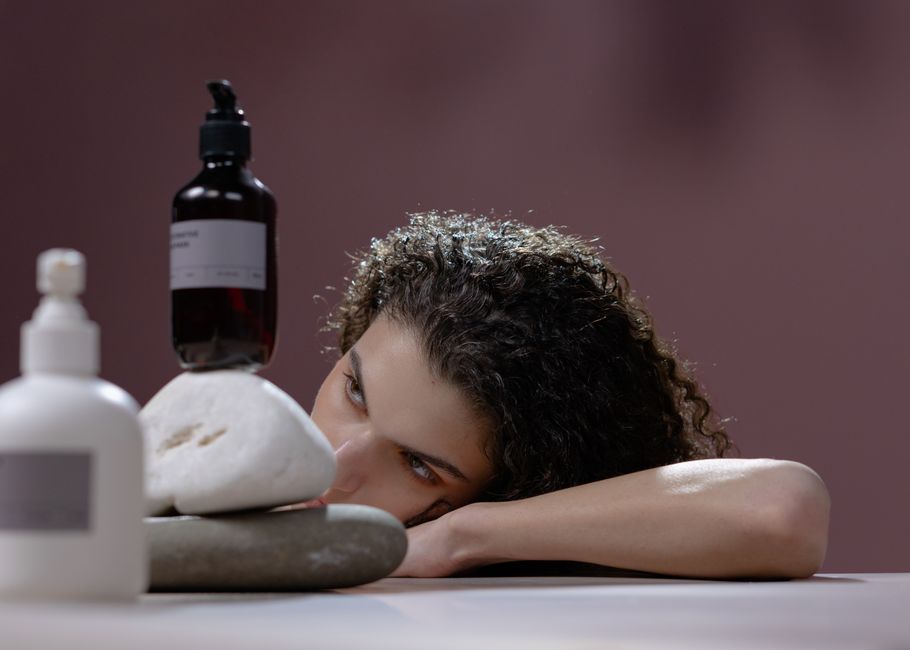 A woman with curly hair leans on a table holding cosmetic bottles atop stacked stones.