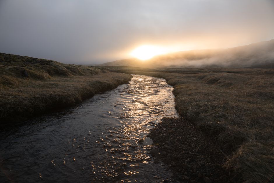Majestic scenery of calm river flowing on rocky riverbed in flat terrain and sun setting down in clouds