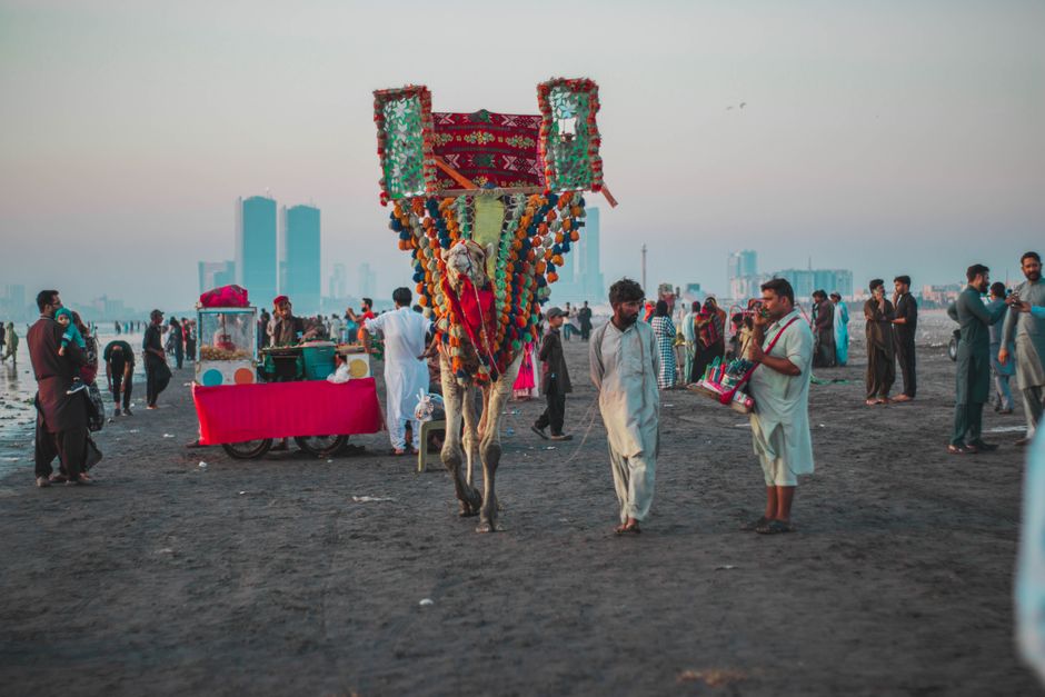Vibrant cultural scene with a decorated camel and crowd on a busy beach with city skyline.