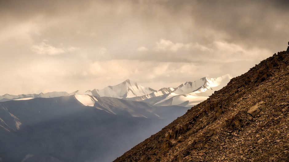 A majestic mountain range with snow-capped peaks bathed in sunlight under a dramatic, cloudy sky.