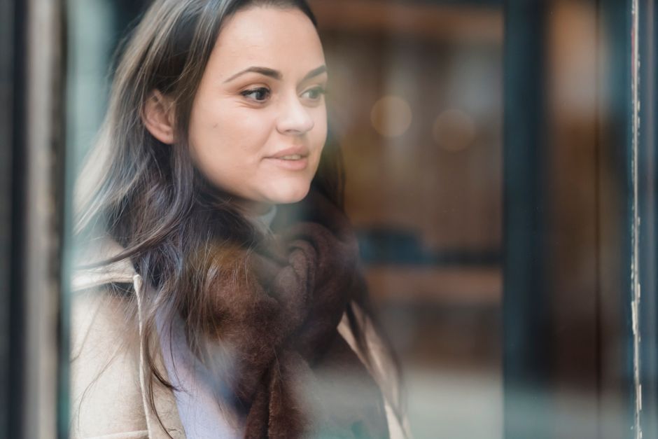 Through glass of crop positive female wearing warm outerwear standing near glass wall and looking away