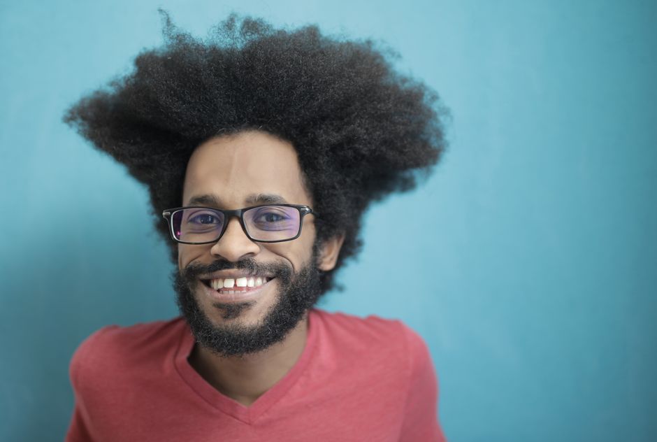 Close-up portrait of a smiling man with a distinctive afro hairstyle and glasses against a blue background.