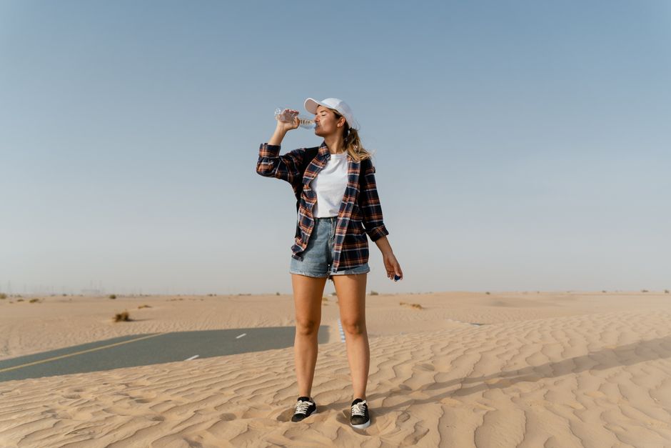 A woman in casual attire drinks water while standing in a vast desert landscape, capturing the essence of travel and adventure.