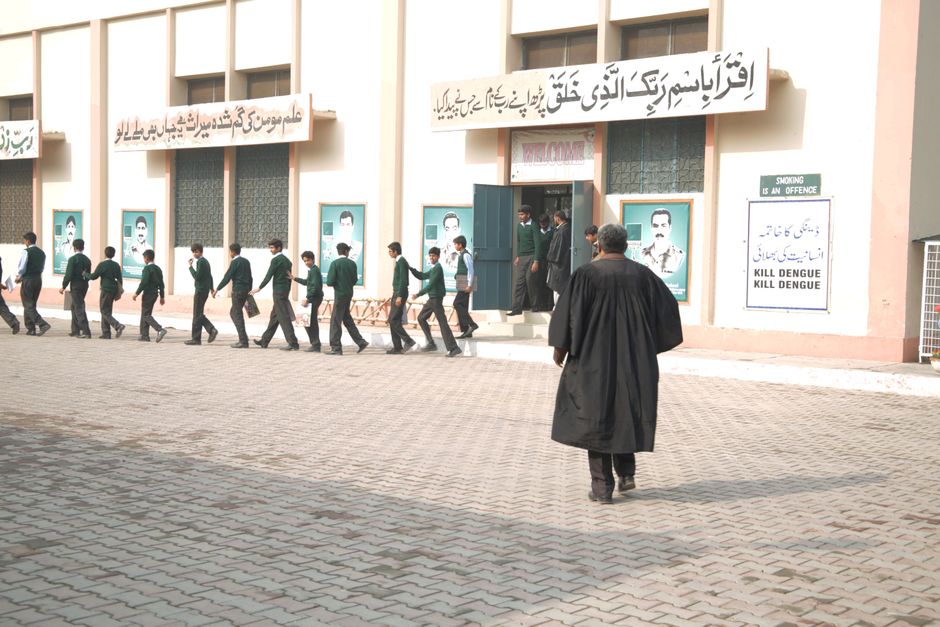 Students in uniform lined up at a school entrance with a teacher overseeing.