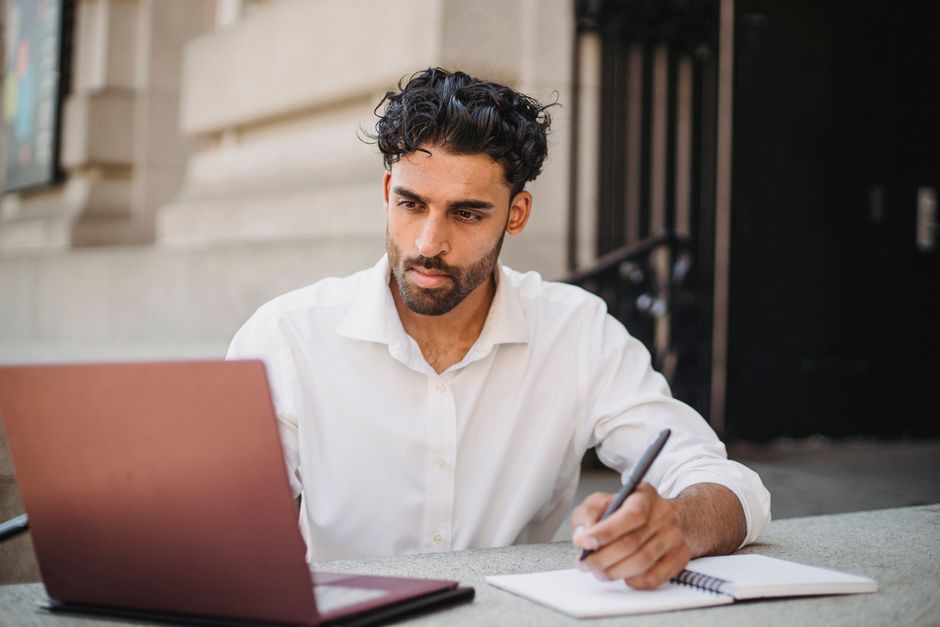 Focused businessman working on laptop outdoors, taking notes in notepad, wearing formal white shirt.