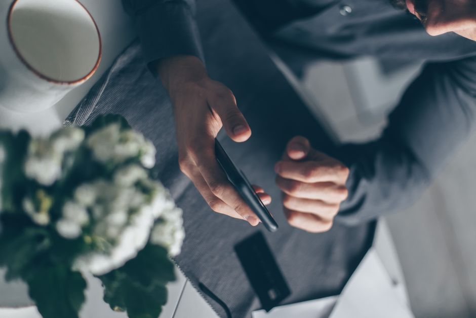 Top view of a man's hands holding a smartphone indoors with a cup and flowers nearby.