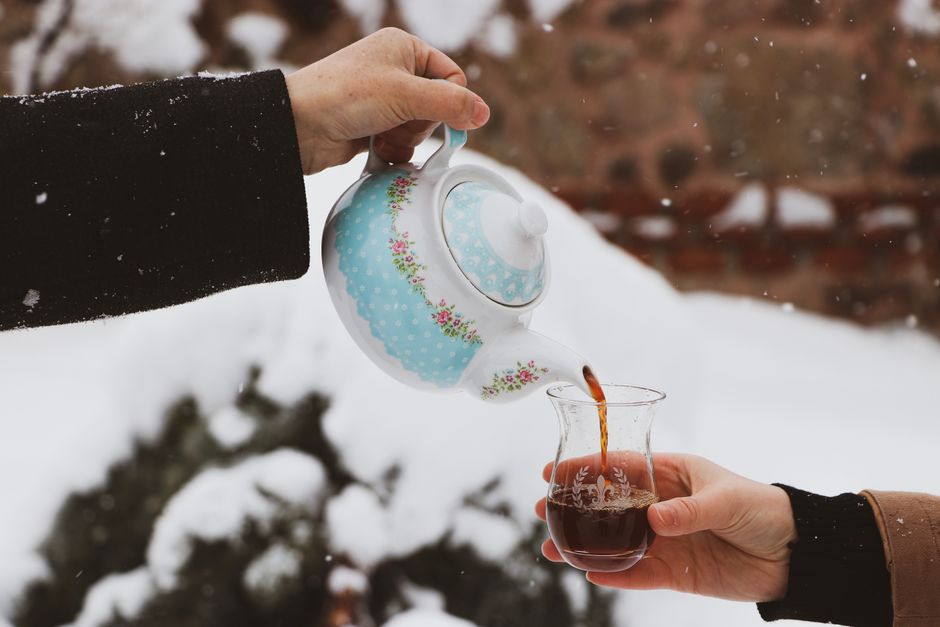 Close-up of hands pouring tea from a floral teapot into a glass in a snowy outdoor scene.