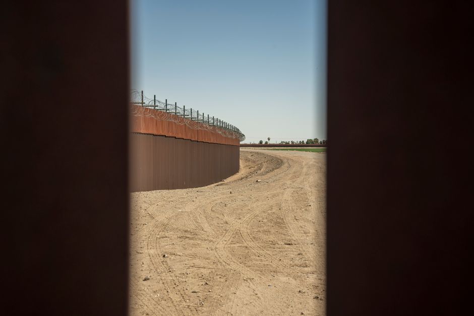 View of a barbed wire border wall structure alongside a dirt road under a clear blue sky.