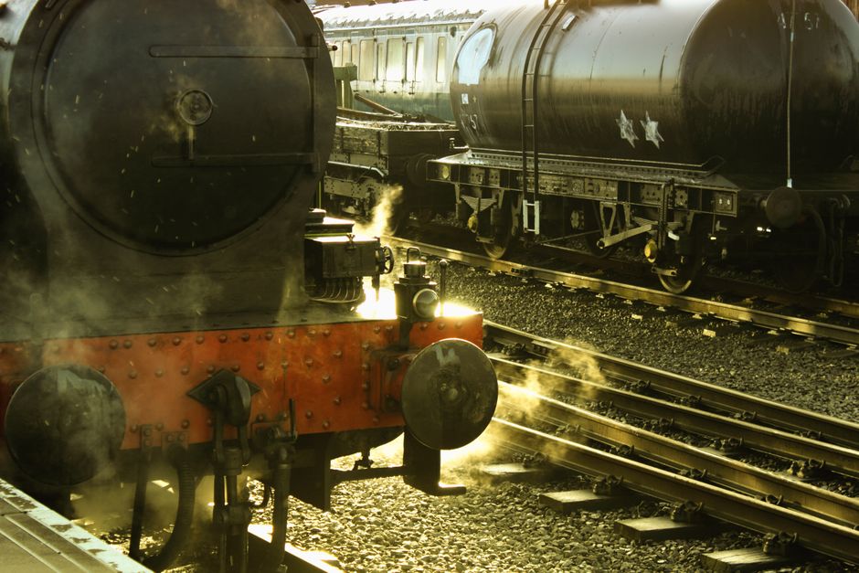 Steam locomotive releasing smoke at an outdoor train station with railroad tracks.