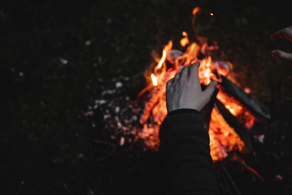 Close-up of hands warming by a campfire, featuring visible flames and burning logs outdoors at night.