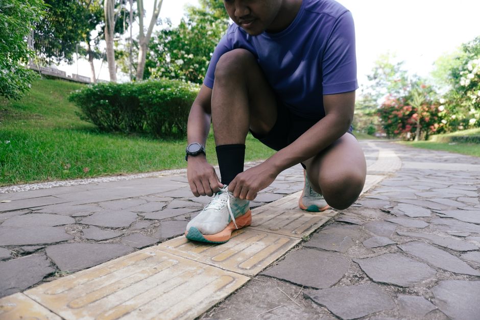 Adult man preparing for a jog by tying his shoelaces on a park trail, surrounded by greenery.