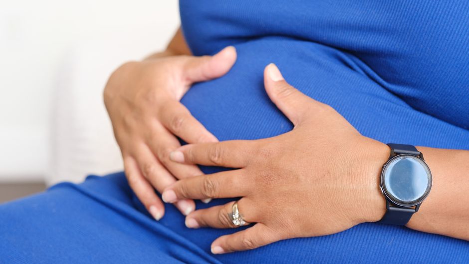 Closeup of a pregnant woman resting her hands on her belly, displaying a watch and a ring.
