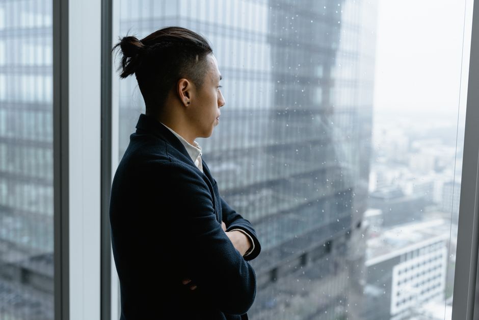 Asian businessman in formal attire looks out from office window on a rainy day.