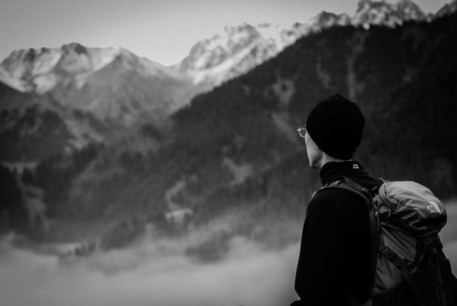 Man with backpack gazes at misty mountain range in black and white.