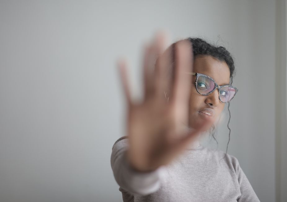 Young African American female wearing casual clothes and eyeglasses outstretching arm at camera while standing near grey wall