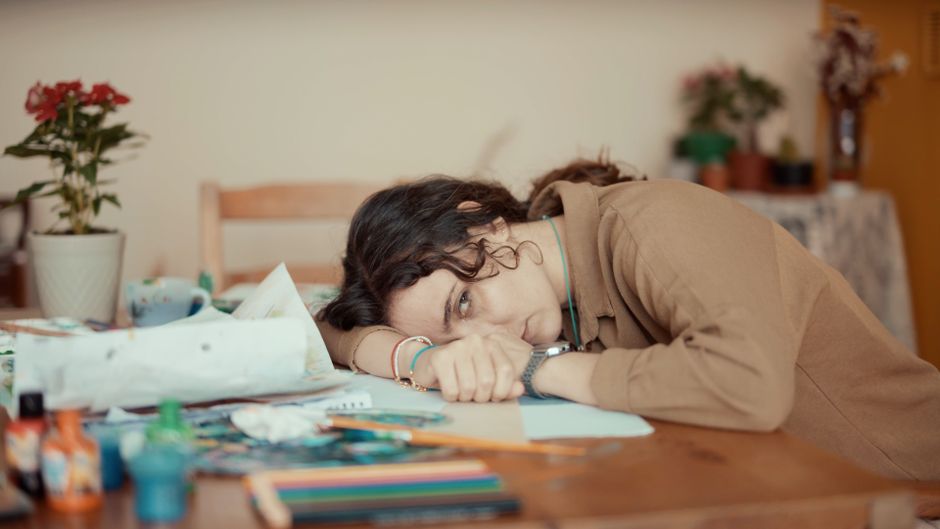 A young woman lying on a desk surrounded by art supplies, appearing tired and thoughtful.
