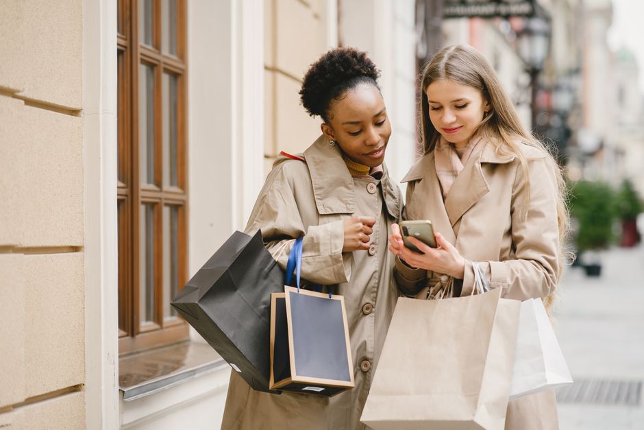 Two women with shopping bags check their phone on a city street.