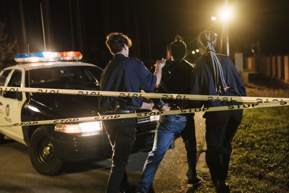 Nighttime arrest scene with police officers and patrol car behind caution tape.