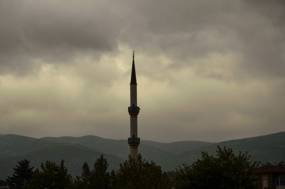 A mosque minaret stands tall against a dramatic cloudy sky with hills in the background.