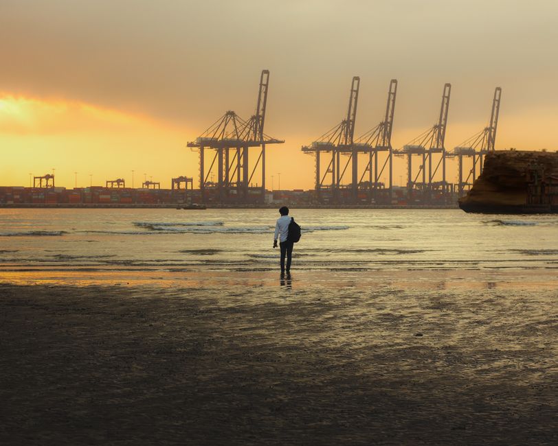 A lone man walks on Karachi beach with port cranes in the background at stunning sunset.