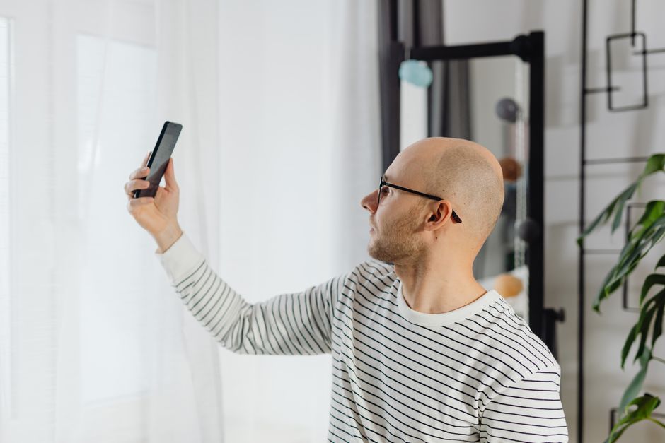 A bald man making a video call using a smartphone indoors, wearing a striped shirt.