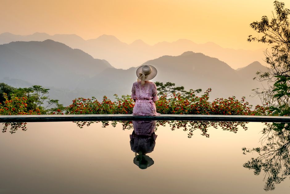 Back view of romantic unrecognizable woman in dress and straw hat sitting on poolside near smooth surface of water and looking away on sunset in mountain terrain