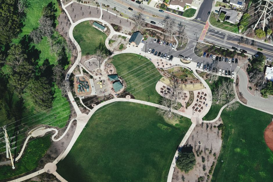 Drone view of modern city park area with winding roads surrounded by greenery and cars parked nearby located under power lines