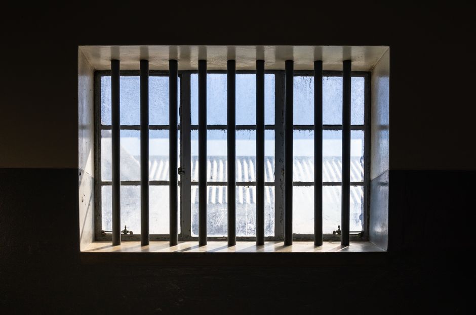 Sunlight streams through barred window of a prison cell, casting shadows and light patterns.