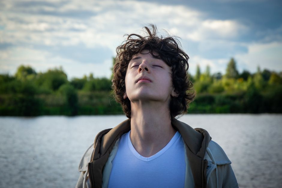 Teen enjoying tranquil moment by the river, eyes closed, in green natural setting.