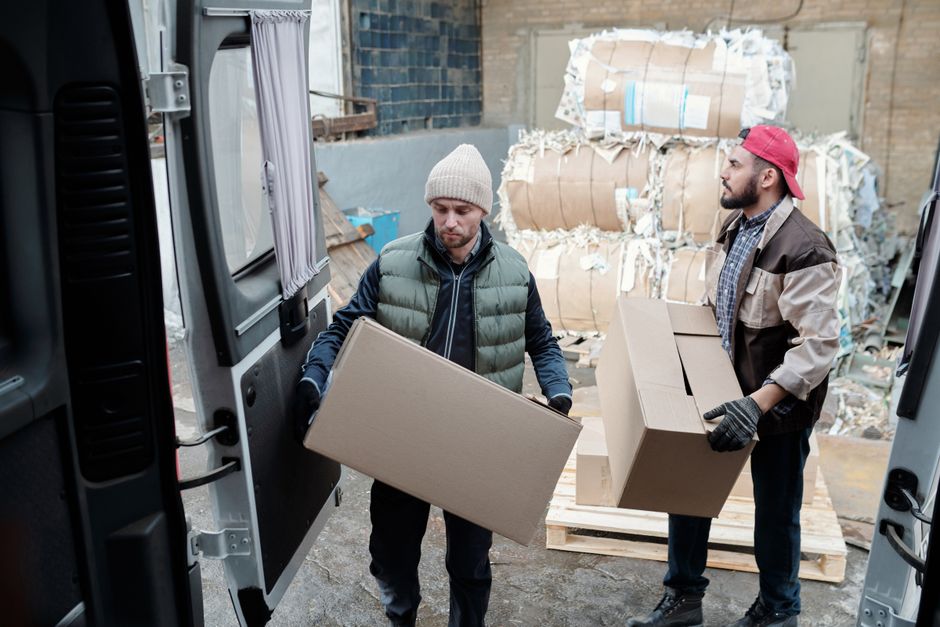 Two male workers loading cardboard boxes in a warehouse with bundled paper in the background.
