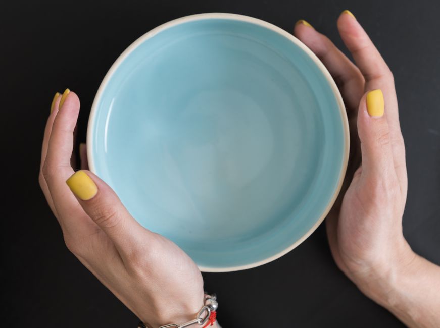 Hands with yellow manicured nails holding a blue ceramic bowl against a dark background.