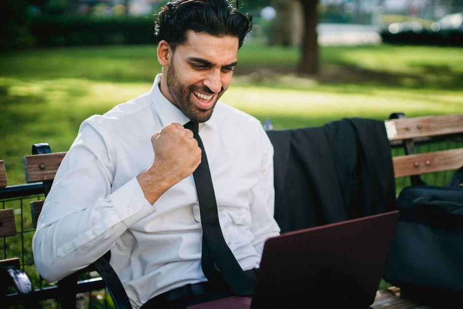 A joyful businessman celebrates a success working on his laptop outdoors on a park bench.