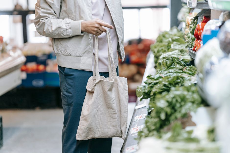 Crop anonymous buyer with shopping bag choosing fresh greens while standing near stall during grocery shopping in supermarket on blurred background