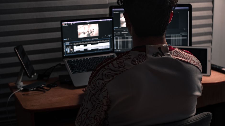 Man sitting at desk working on video editing software with dual monitors in an office.