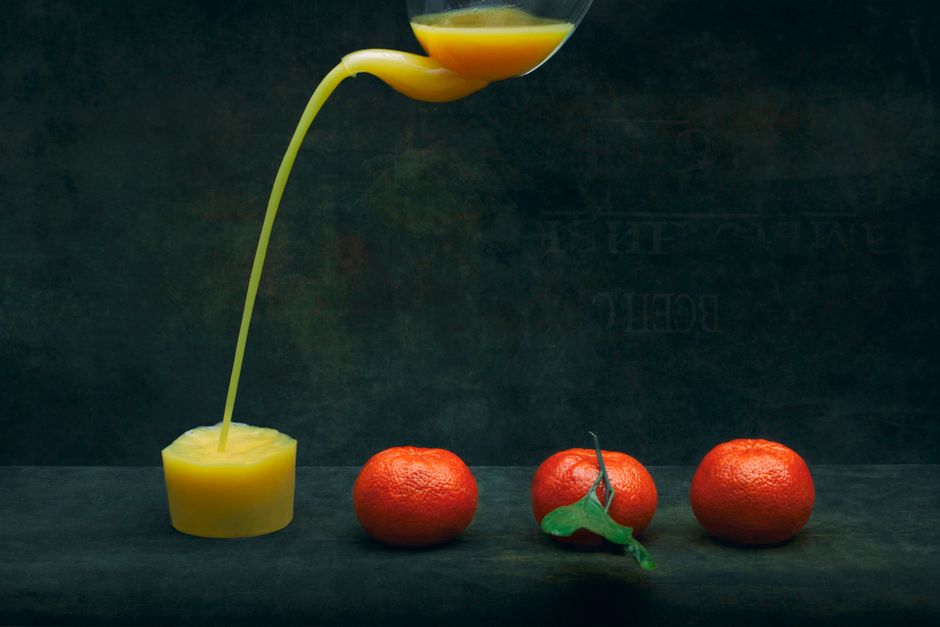 Artistic shot of fresh orange juice pouring from a glass with three oranges on a dark backdrop.