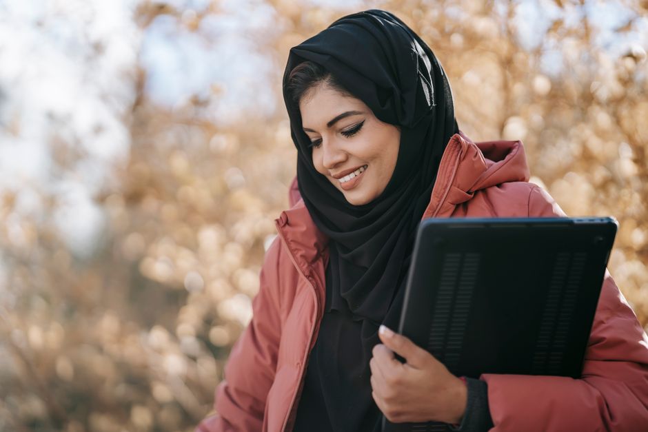 Smiling Muslim woman freelancer in black headscarf standing with netbook in hand and looking down on street against tree with autumn foliage in daylight