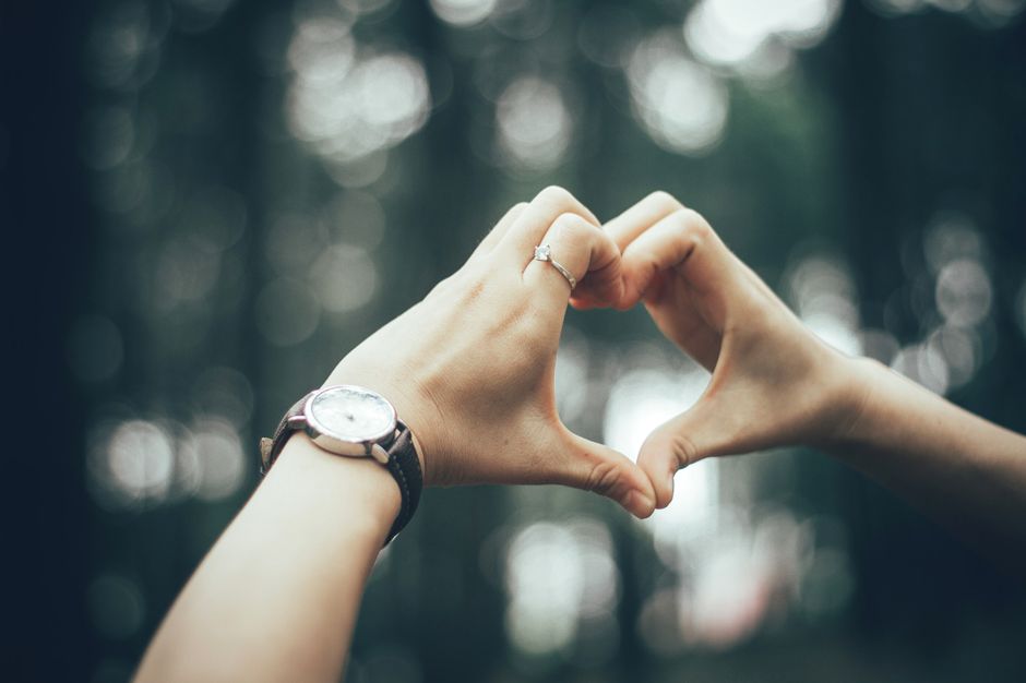 A pair of hands forming a heart shape with a focus on a ring and wristwatch, outdoors.