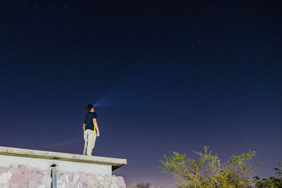 A person on a rooftop uses a flashlight to illuminate the starry night sky.