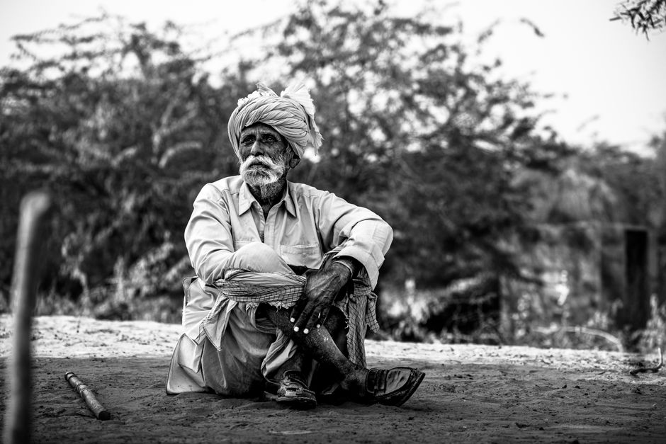 A thoughtful portrait of an elderly man in a turban sitting outdoors, depicted in black and white.