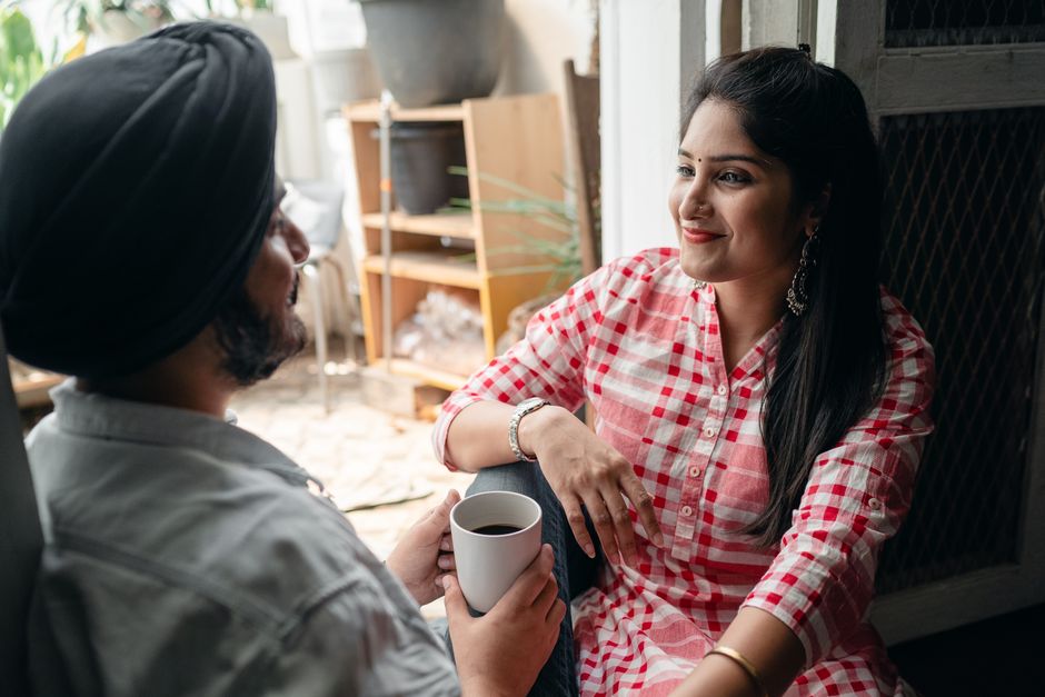 Cheerful young wife with bindi on forehead wearing plaid dress sitting in doorway against street kitchen with young bearded husband in turban while communicating and drinking tea and looking at each other