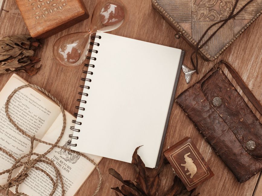 A vintage-style flatlay with an open notebook, book, hourglass, and leather items on a wooden surface.