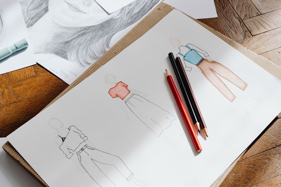 Artistic fashion design sketches on paper accompanied by colored pencils on a wooden surface.