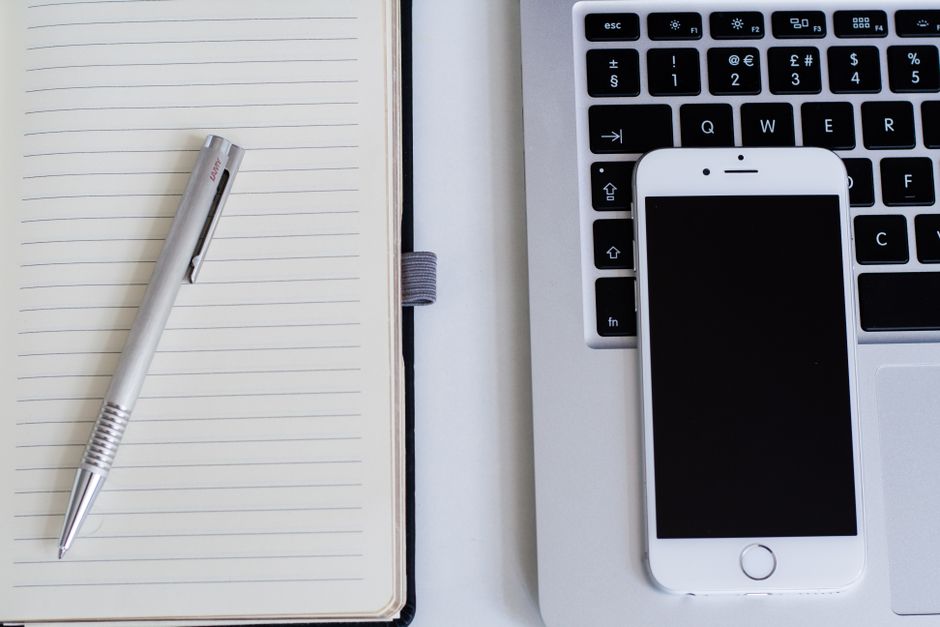 Close-up of a notebook, pen, smartphone, and laptop on a modern workspace.