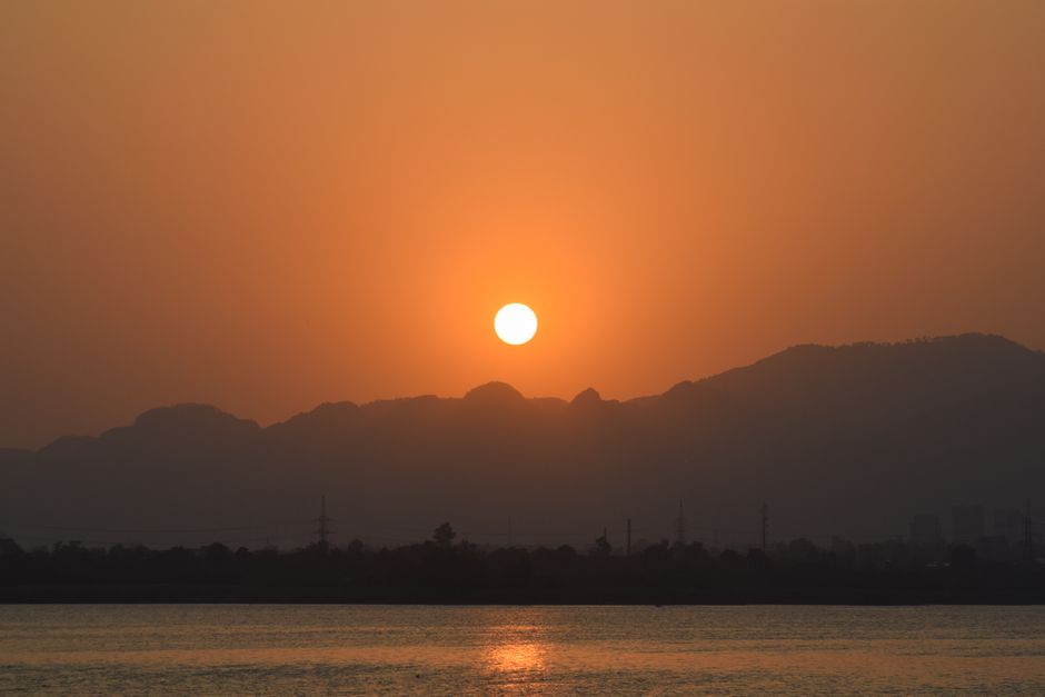 Peaceful sunset view with sun setting over distant mountains and calm water surface.