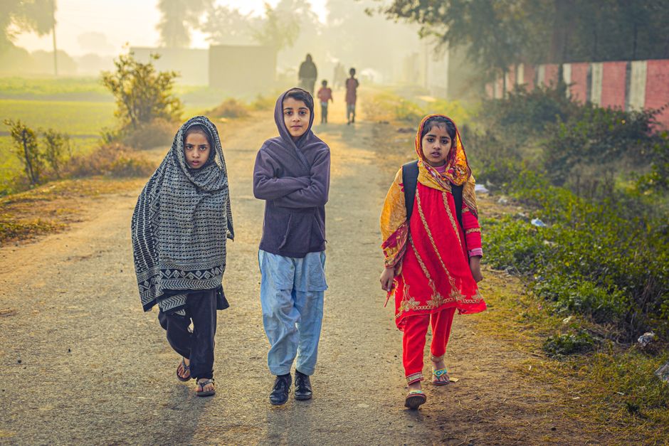 Three children stroll along a rural path in Pakistan during daytime, capturing a sense of community and tradition.