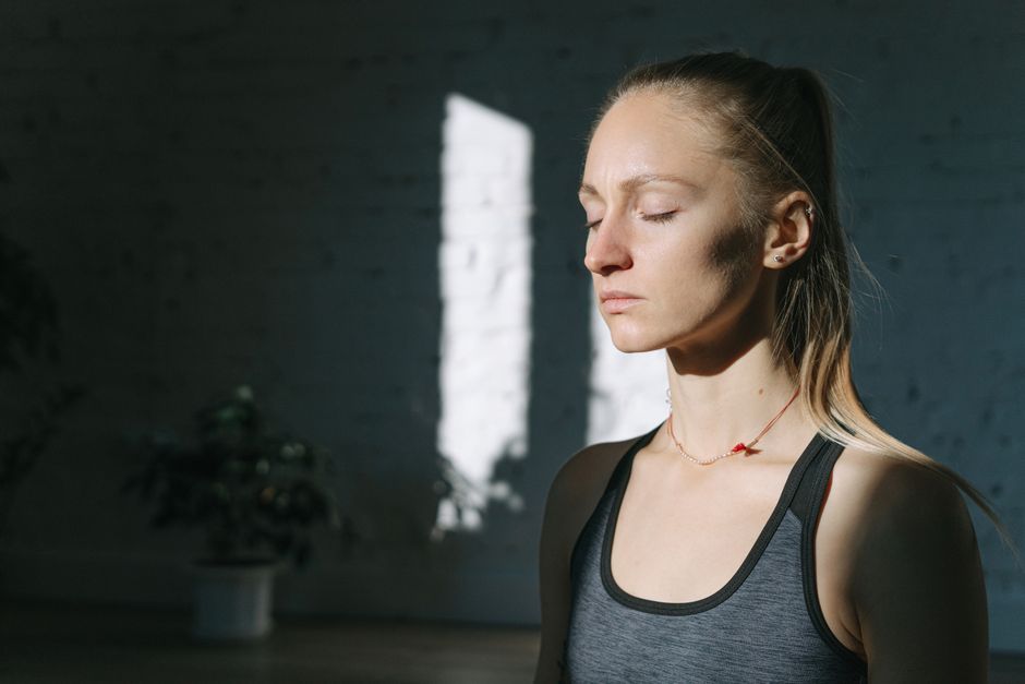 A woman in meditation inside a sunlit room, eyes closed, feeling calm and peaceful.