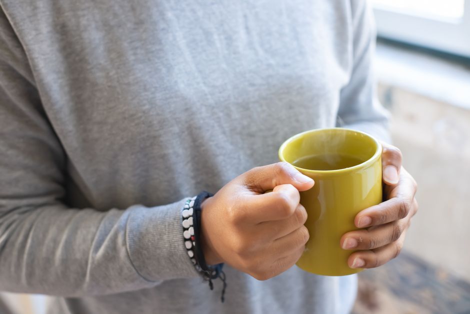 Close-up of individual in gray sweater holding a steaming mug of hot tea indoors.