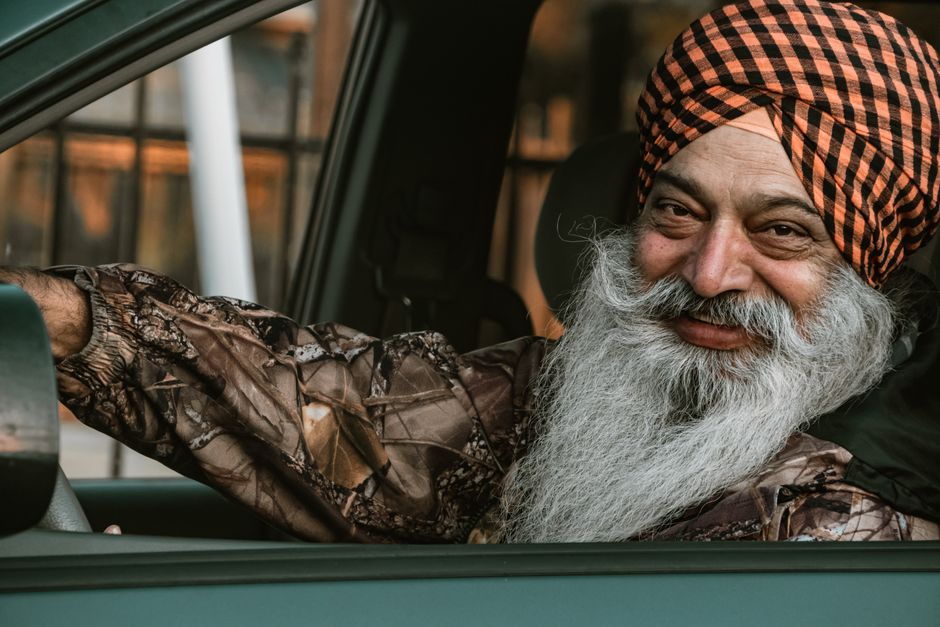 Elderly man with a beard and turban smiling while sitting in a car.